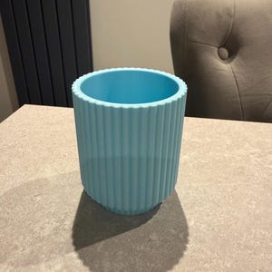 May include: A light blue cylindrical container with vertical ribbed detailing. The container is made of a solid material and sits on a neutral-toned surface. The container's shadow is visible on the surface.