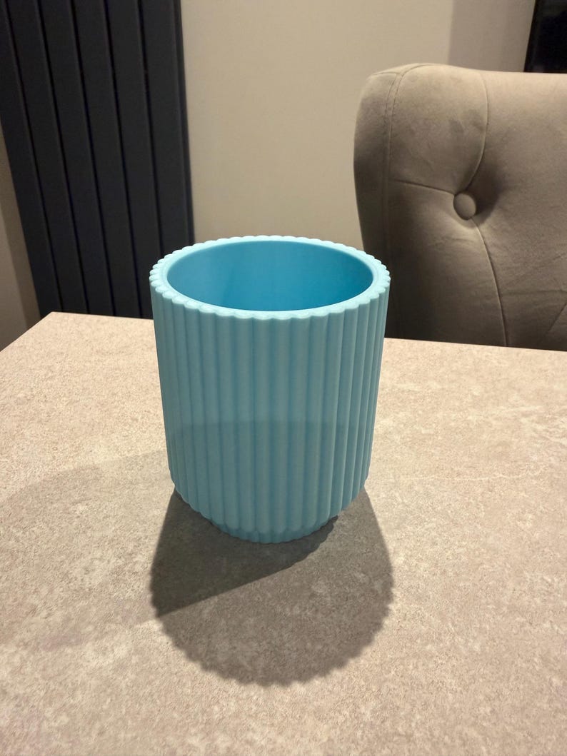 May include: A light blue cylindrical container with vertical ribbed detailing. The container is made of a solid material and sits on a neutral-toned surface. The container's shadow is visible on the surface.