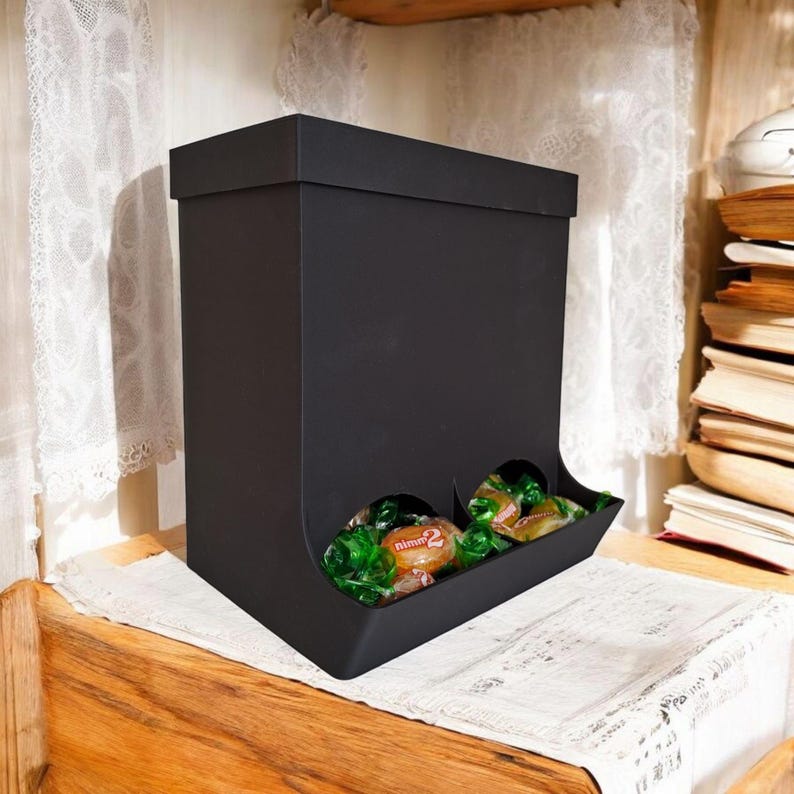 May include: A black candy dispenser with a rectangular shape and a lid. The dispenser has two openings at the bottom, each filled with wrapped candies. The candies are green and brown. The dispenser is on a white lace cloth.