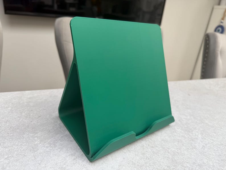 May include: A vibrant green tablet stand. The stand has a triangular base and a slightly angled back, with a small lip at the bottom to hold a tablet. The stand is made of a solid material and is designed to hold a tablet upright.