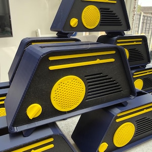May include: A collection of navy blue and yellow geometric boxes. Each box features a yellow circular speaker and a yellow knob. The boxes are stacked, showcasing their design and color scheme. The overall aesthetic is modern and industrial.