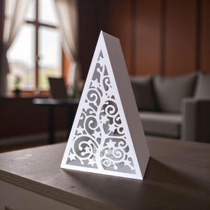 May include: A white, pyramid-shaped decorative light with intricate cut-out designs. The cut-outs form a tree-like pattern, creating a play of light and shadow. The lamp sits on a wooden surface, with a blurred background.