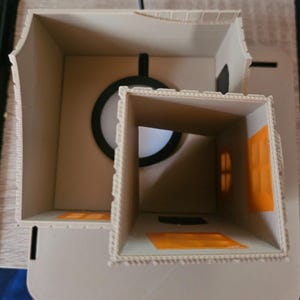 May include: A beige, multi-room model house with a circular window and rectangular openings. The model has orange accents and is constructed with a textured material. The design includes a chimney-like structure and a flat base.