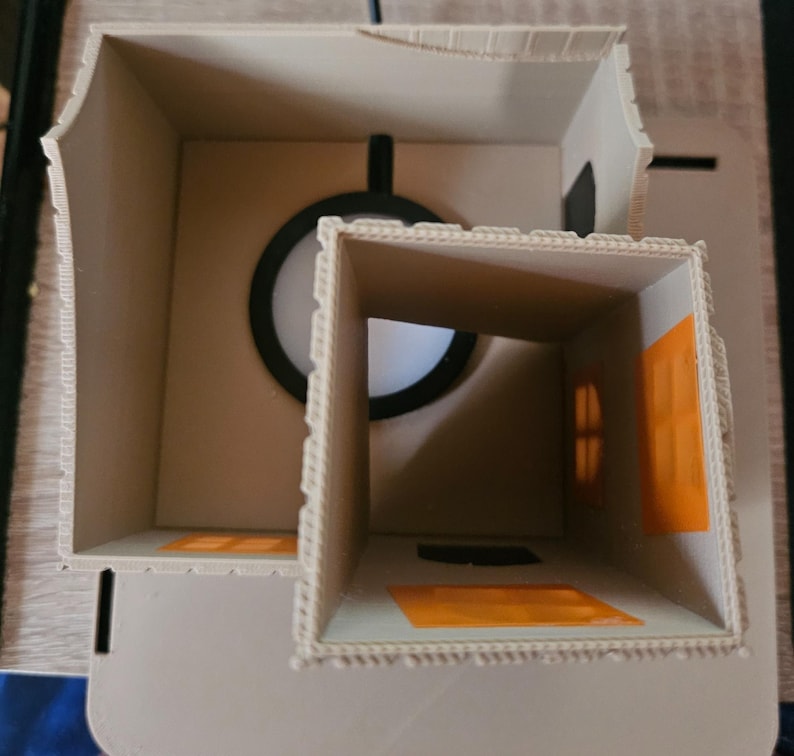 May include: A beige, multi-room model house with a circular window and rectangular openings. The model has orange accents and is constructed with a textured material. The design includes a chimney-like structure and a flat base.
