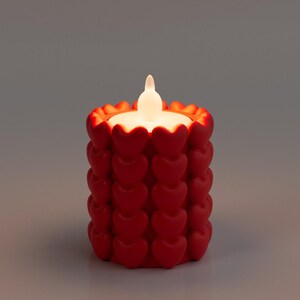 May include: A red candle with a heart-shaped design. The candle has a white, flickering LED light. The candle is cylindrical and sits on a gray surface. The candle is a decorative item.