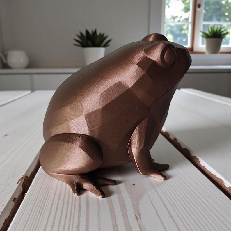 May include: A bronze-colored, geometric frog sculpture. The frog is posed in a sitting position with its front legs extended. The sculpture has a faceted design, giving it a modern look. The frog is sitting on a white wooden surface.