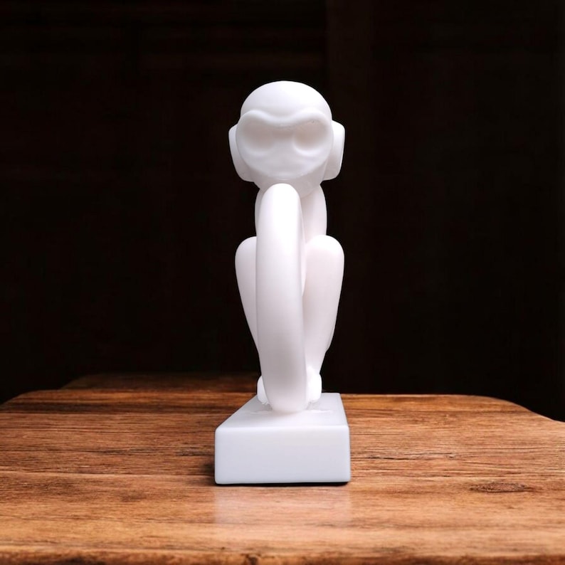 Puede incluir: Una figura de mono blanco, sentado con las piernas recogidas, se exhibe sobre una base rectangular. La escultura tiene un acabado liso y mate y se coloca sobre un fondo oscuro. Los rasgos del mono est&aacute;n estilizados, con orejas grandes y una expresi&oacute;n pensativa.