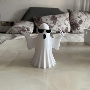 May include: A white ghost figurine wearing black sunglasses and gesturing with its hands. The ghost has a simple, cartoonish design and is standing on a light-colored surface. The background includes a bed with floral pillows.