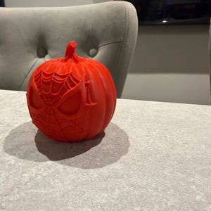 May include: A bright red pumpkin with a Spider-Man face design carved into the surface. The pumpkin is sitting on a gray surface. The pumpkin is a festive decoration for Halloween.