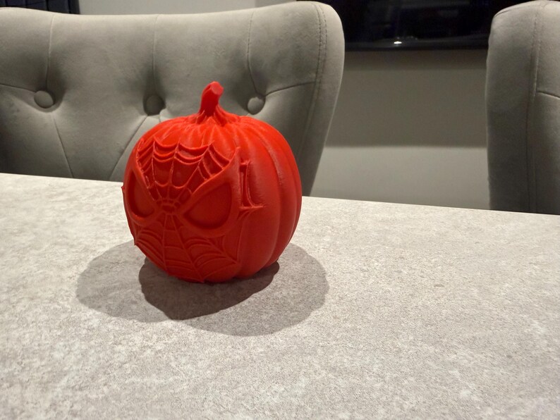 May include: A bright red pumpkin with a Spider-Man face design carved into the surface. The pumpkin is sitting on a gray surface. The pumpkin is a festive decoration for Halloween.