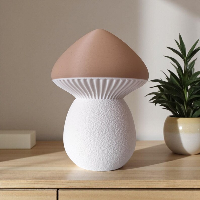 May include: A mushroom-shaped lamp with a textured white base and a light brown cap. The lamp has a ribbed, white underside and sits on a wooden surface. A small potted plant is visible in the background.