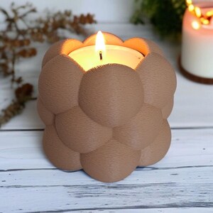 May include: A lit candle in a brown, bubble-shaped holder. The candle has a white wax base and a flickering flame. The holder is made of a textured material and sits on a light-colored wooden surface.