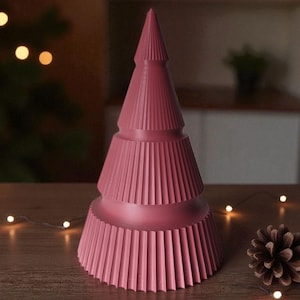 May include: A pink, stylized Christmas tree decoration with a ribbed design. The tree is on a wooden surface, with a pine cone and string lights nearby. The tree has a conical shape with layered sections.