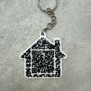 May include: White house-shaped keychain with a black QR code design. The keychain has a silver chain and a keyring. The house shape has a chimney and a roof. The QR code is made of black squares and dots.