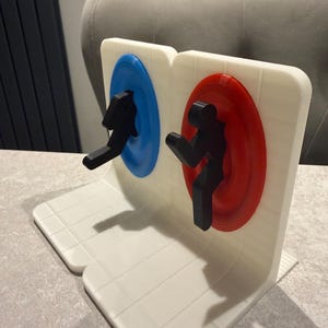 May include: A white, 3D-printed bookend featuring two circular portals, one blue and one red, with black silhouettes of figures. The bookend has a grid-like pattern on the base and is designed to hold books upright.