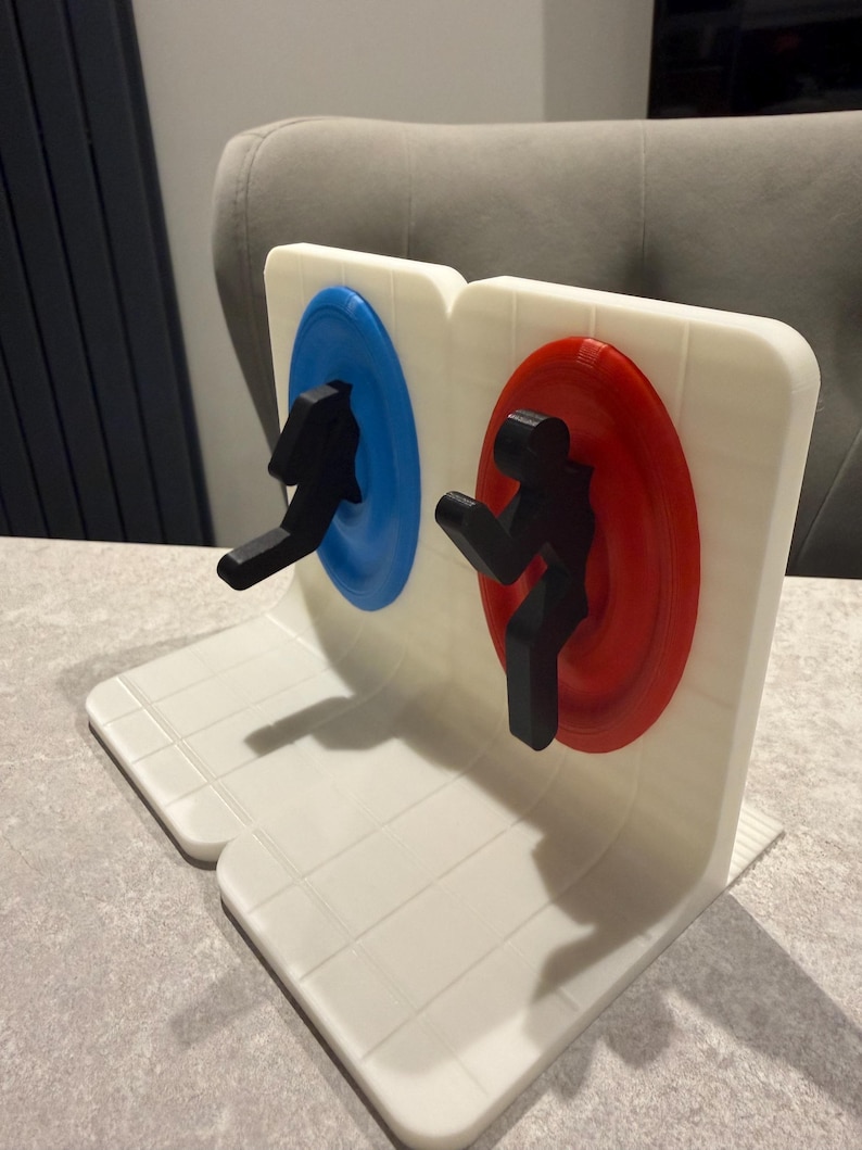 May include: A white, 3D-printed bookend featuring two circular portals, one blue and one red, with black silhouettes of figures. The bookend has a grid-like pattern on the base and is designed to hold books upright.