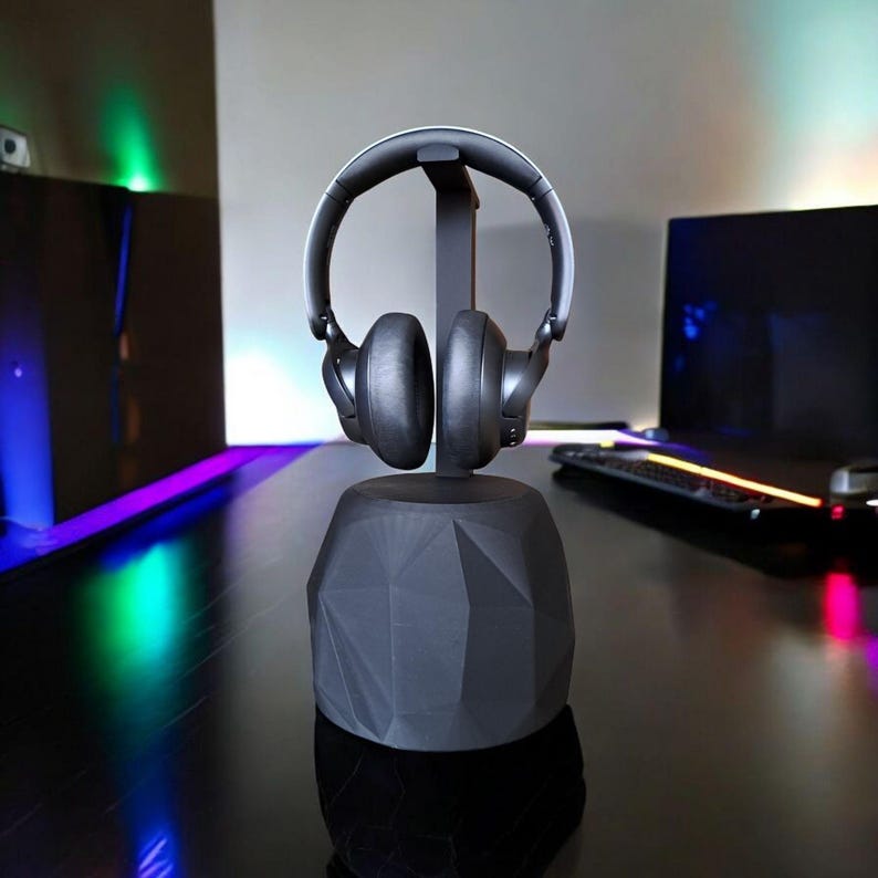 May include: A black headphone stand with a geometric design, holding a pair of black headphones. The stand has a faceted base and a vertical support for the headphones. The headphones are black with a silver band.