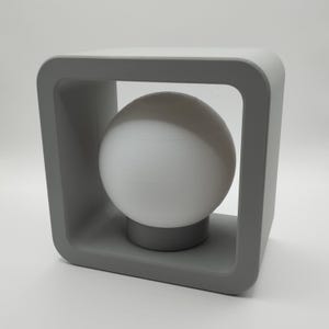 May include: A modern gray table lamp with a spherical white light bulb. The lamp features a square frame design with rounded corners, and the bulb rests on a small, circular base. The lamp is a neutral color and suitable for any room.