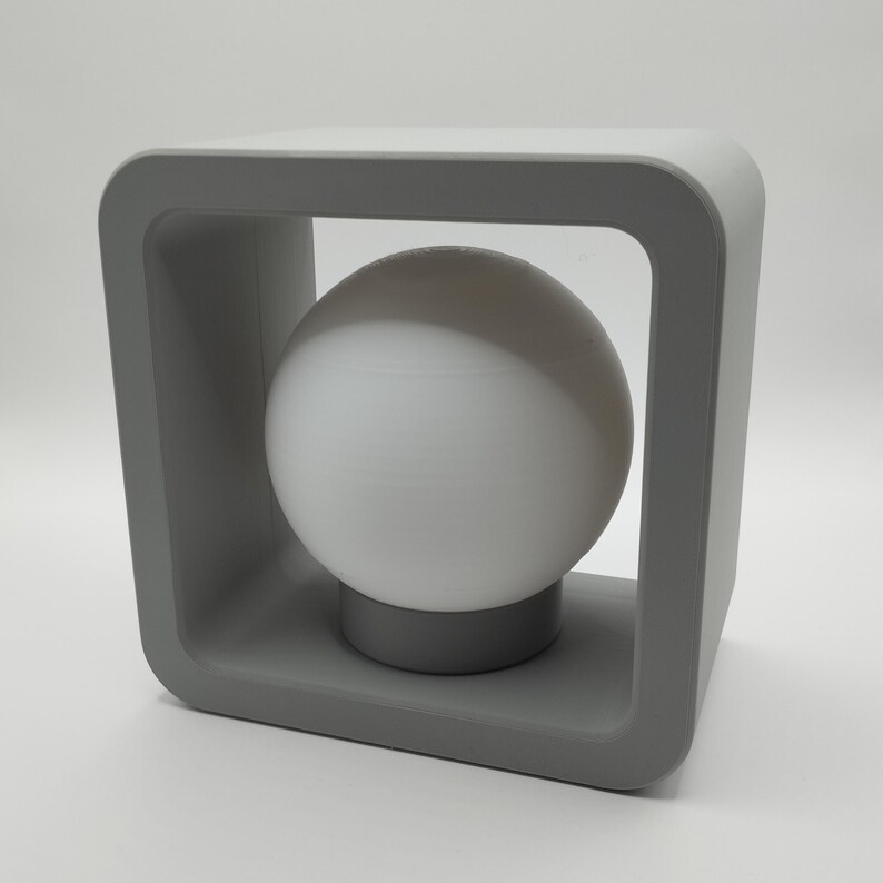 May include: A modern gray table lamp with a spherical white light bulb. The lamp features a square frame design with rounded corners, and the bulb rests on a small, circular base. The lamp is a neutral color and suitable for any room.