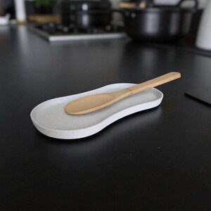 May include: A light grey speckled spoon rest with a wooden spoon. The spoon rest has a curved, organic shape. The background is a black countertop with kitchen items out of focus.