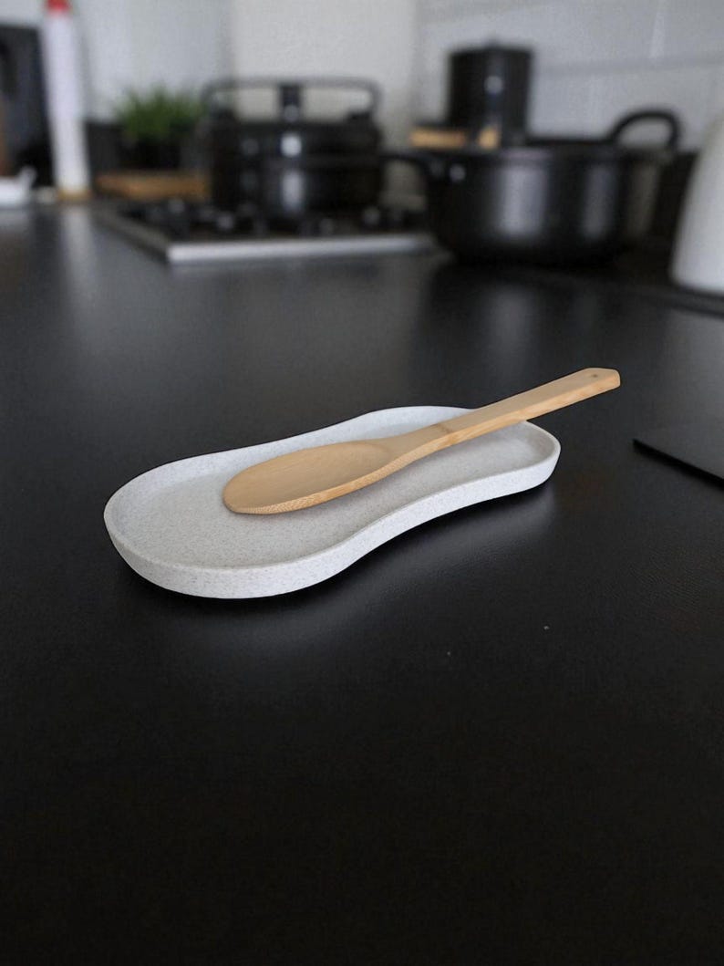 May include: A light grey speckled spoon rest with a wooden spoon. The spoon rest has a curved, organic shape. The background is a black countertop with kitchen items out of focus.