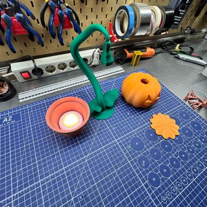 May include: A green, 3D-printed lamp with a curved stem and a small lamp head, next to an orange pumpkin with carved details. A small orange flower and a candle in a terracotta pot are also visible.