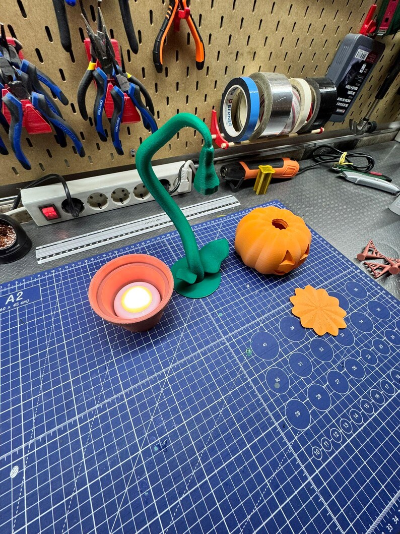 May include: A green, 3D-printed lamp with a curved stem and a small lamp head, next to an orange pumpkin with carved details. A small orange flower and a candle in a terracotta pot are also visible.