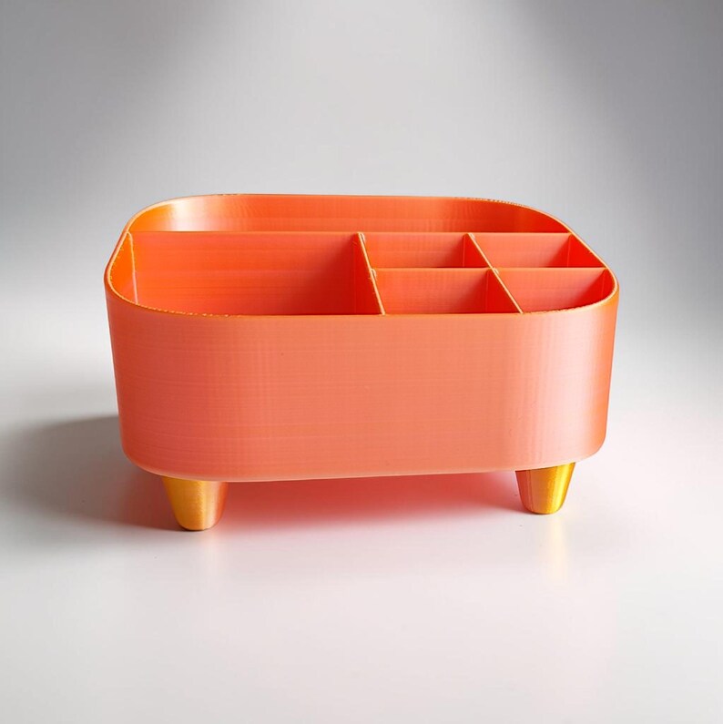 May include: A coral-colored organizer with six compartments, resting on three gold-colored, rounded legs. The organizer is made of a solid material and is designed to hold small items.