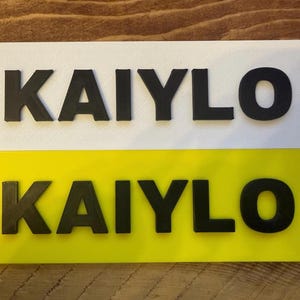 Pu&ograve; includere: Due targhe rettangolari, una bianca e una gialla, che mostrano il nome "KAIYLO" in lettere nere e in grassetto. Le targhe sono probabilmente per un veicolo o un oggetto personale.