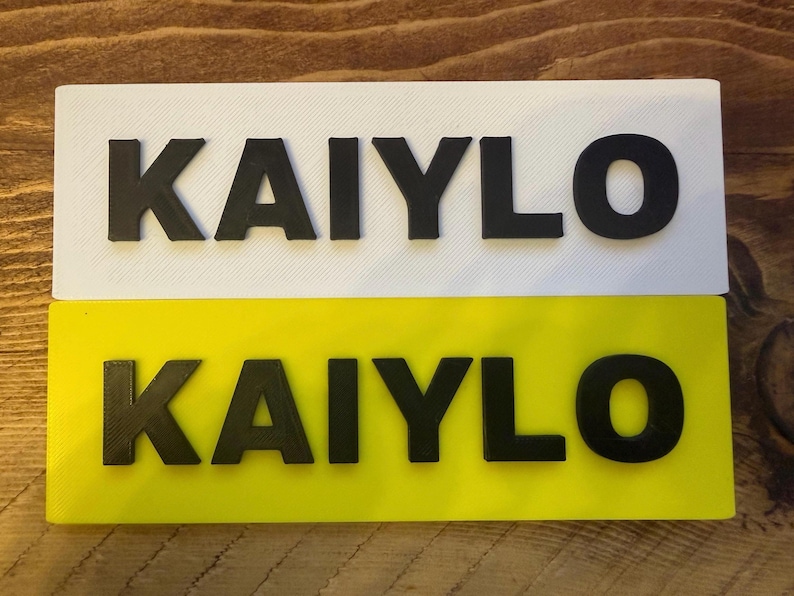 Pu&ograve; includere: Due targhe rettangolari, una bianca e una gialla, che mostrano il nome "KAIYLO" in lettere nere e in grassetto. Le targhe sono probabilmente per un veicolo o un oggetto personale.