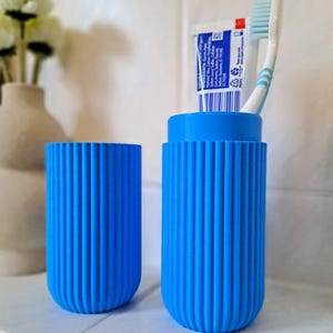 May include: Two bright blue, ribbed toothbrush holders. One holder contains a tube of toothpaste and a toothbrush. The holders are cylindrical with rounded bottoms. The background includes a white vase with flowers.