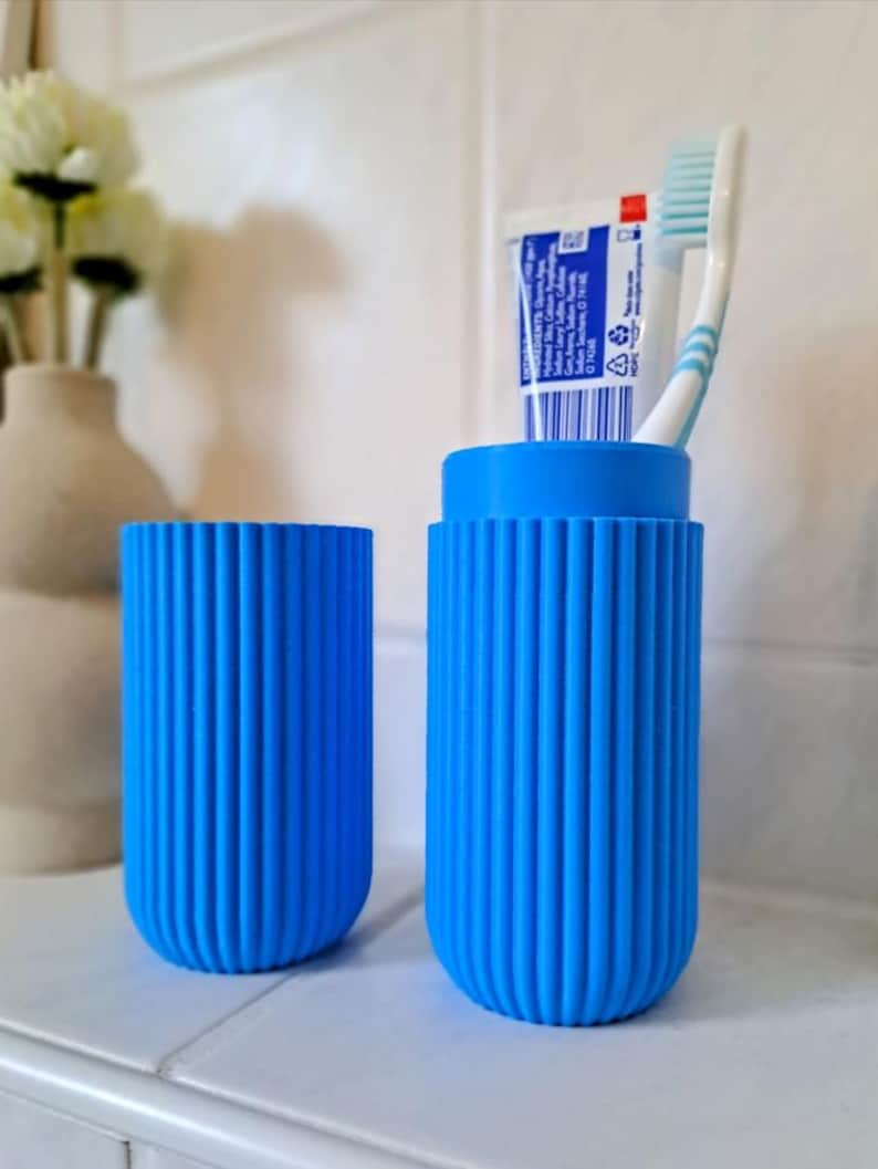 May include: Two bright blue, ribbed toothbrush holders. One holder contains a tube of toothpaste and a toothbrush. The holders are cylindrical with rounded bottoms. The background includes a white vase with flowers.