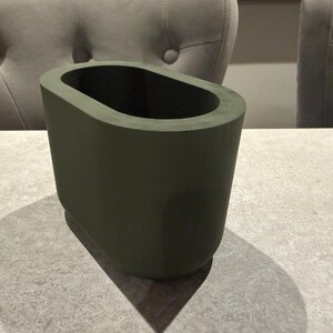 May include: An olive green, oval-shaped vase. The vase has a smooth, matte finish and a rectangular base. The interior is dark, creating a contrast with the exterior. The vase is sitting on a light gray surface.