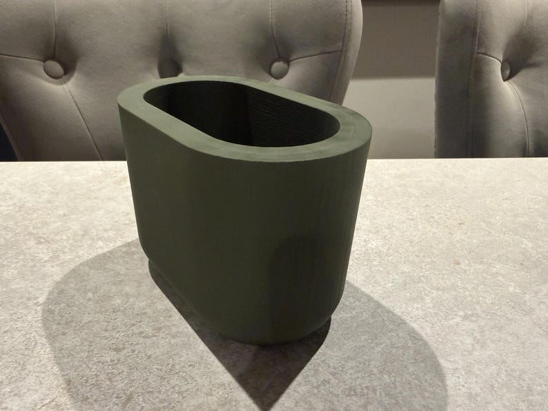 May include: An olive green, oval-shaped vase. The vase has a smooth, matte finish and a rectangular base. The interior is dark, creating a contrast with the exterior. The vase is sitting on a light gray surface.