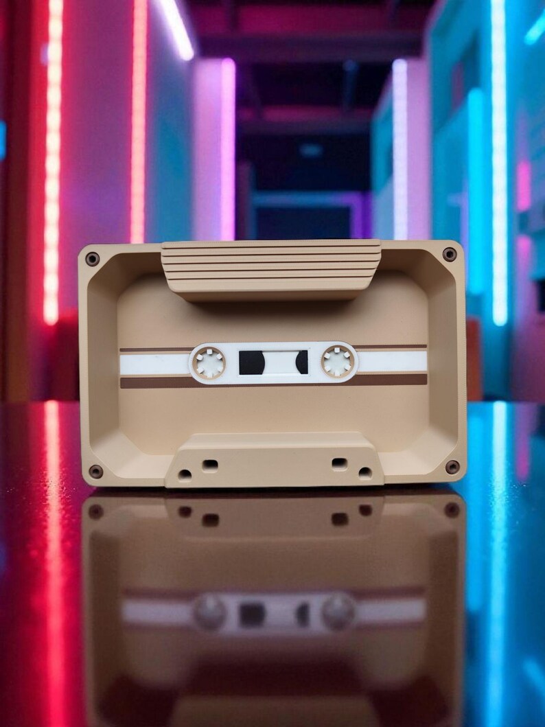 Pu&ograve; includere: Un contenitore beige a forma di musicassetta con dettagli bianchi e marroni. Il contenitore ha una forma rettangolare con angoli arrotondati e piccoli dettagli circolari. Il design imita una classica musicassetta.
