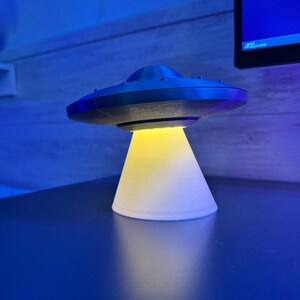 May include: A 3D-printed UFO lamp with a black top and a white base. The lamp emits a yellow light from the bottom, creating a glowing effect. The lamp is sitting on a dark blue surface.