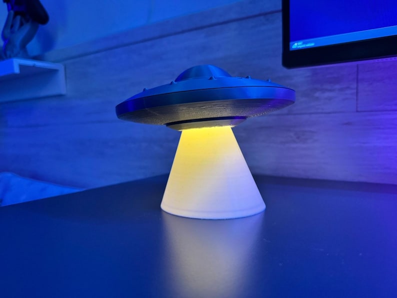 May include: A 3D-printed UFO lamp with a black top and a white base. The lamp emits a yellow light from the bottom, creating a glowing effect. The lamp is sitting on a dark blue surface.