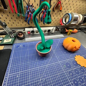 May include: A green, stylized plant in a small brown pot, with a curved stem and a flower-like bud. The plant sits on a blue cutting mat, next to an orange pumpkin and a leaf-shaped cutout. Various tools hang on the wall in the background.