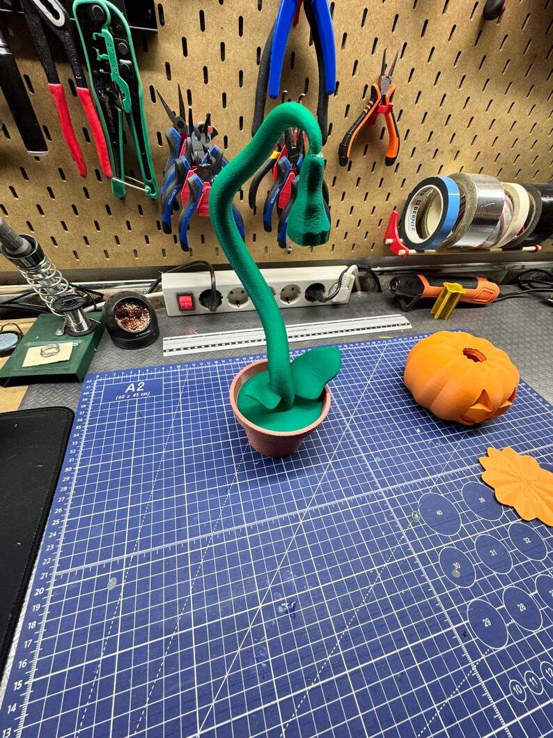 May include: A green, stylized plant in a small brown pot, with a curved stem and a flower-like bud. The plant sits on a blue cutting mat, next to an orange pumpkin and a leaf-shaped cutout. Various tools hang on the wall in the background.