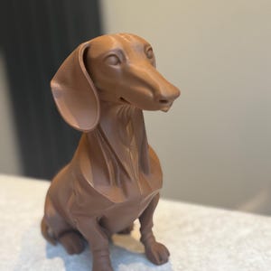 May include: A brown dachshund dog figurine. The dog is sitting upright with its ears flopped down. The figurine is a solid color and has a smooth texture. The dog's features are well-defined, including its long snout and paws.