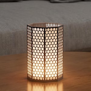 Op de afbeelding: Een decoratieve, zeshoekige lantaarn met een honingraatpatroon. Het zwarte metalen frame omsluit een warm, gloeiend licht, waardoor een zachte sfeer ontstaat. De lantaarn staat op een houten oppervlak, wat wijst op een gezellige interieursetting.