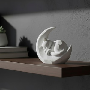 Peut inclure: Une figurine de chat blanc dormant paisiblement dans un croissant de lune. La sculpture est pos&eacute;e sur une &eacute;tag&egrave;re marron fonc&eacute; contre un mur gris. Le chat est enroul&eacute;, et la lune a une surface textur&eacute;e.