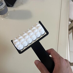 May include: A black handheld massage roller with a white textured surface. The roller is designed with a series of rounded, raised bumps. The handle is black and rectangular, providing a firm grip for use.