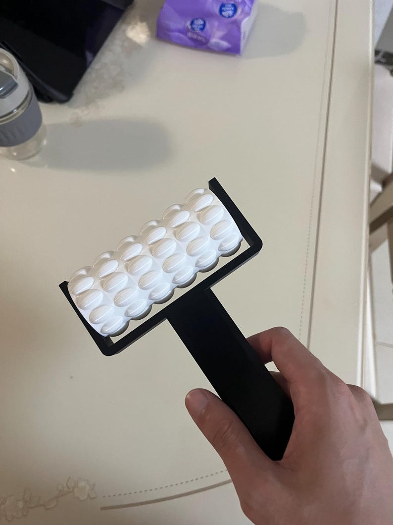 May include: A black handheld massage roller with a white textured surface. The roller is designed with a series of rounded, raised bumps. The handle is black and rectangular, providing a firm grip for use.