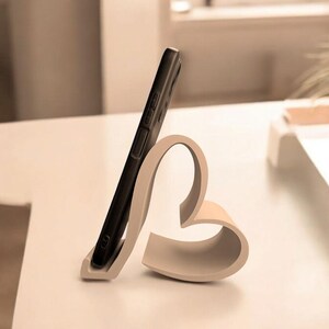 May include: A white heart-shaped phone stand with a black smartphone resting in it. The stand is on a white surface.