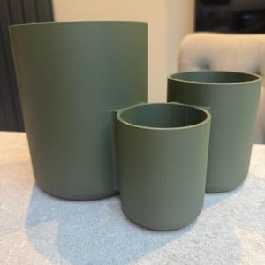 May include: Three olive green cylindrical containers of varying sizes. The containers are made of a solid material and have a smooth, matte finish. The largest container is approximately 6 inches tall, while the smallest is about 3 inches tall.