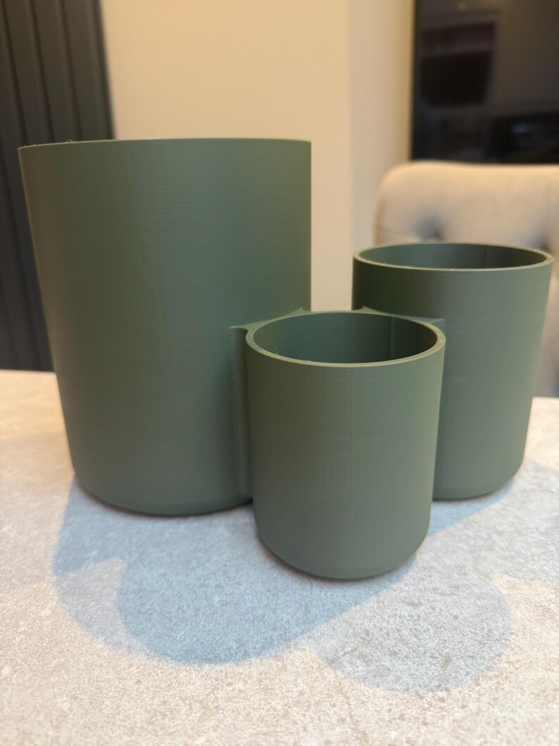 May include: Three olive green cylindrical containers of varying sizes. The containers are made of a solid material and have a smooth, matte finish. The largest container is approximately 6 inches tall, while the smallest is about 3 inches tall.