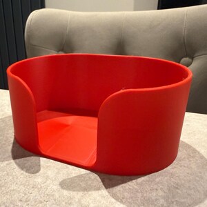 May include: A bright red, rectangular container with rounded corners. The container has a low profile and is open at the top, with a curved cutout on each side. The container is sitting on a light gray surface.