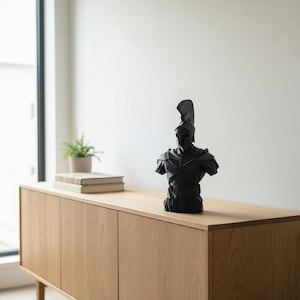 May include: A black sculpture of a Spartan warrior, featuring a helmet and armour. The bust is placed on a light wood cabinet. A small plant and books are visible in the background. A decorative item.