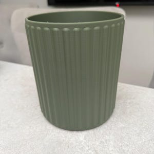 May include: A sage green cylindrical planter with vertical ribbed detailing. The planter has a slightly flared top and a smooth, rounded base. The color is a muted, earthy tone, suitable for various home decor styles. The planter is likely made of plastic or a similar material.